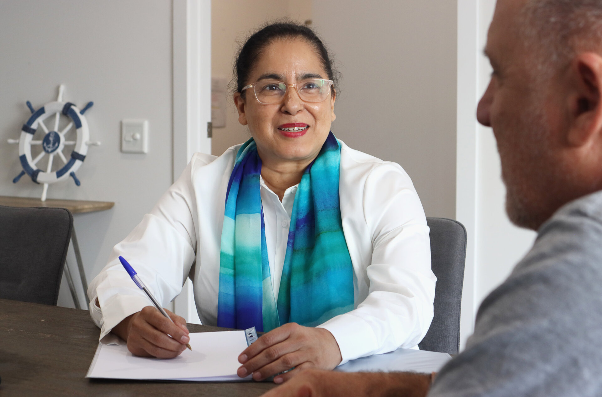 A woman sitting at a desk writing on some paper while talking to a man, in profile.